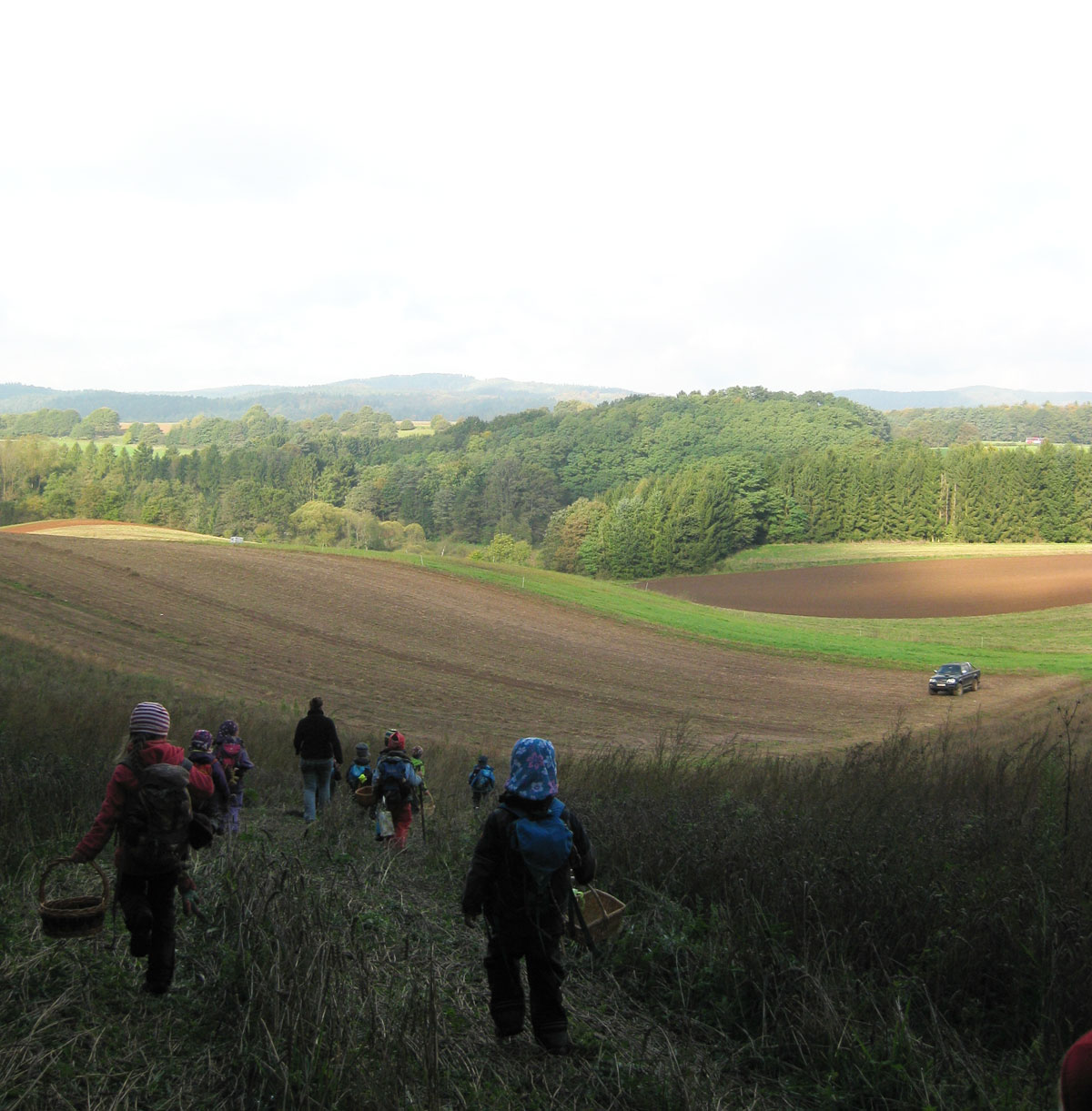 Waldkindergarten-Merzig-Besseringen Von einem Hügel aus geht es tief ein Tal mit vielen Feldern. Am Horizont ist der Wald zu sehen. Einige Kindern rennen oder gehen den Hügel zum Tal hinunter.